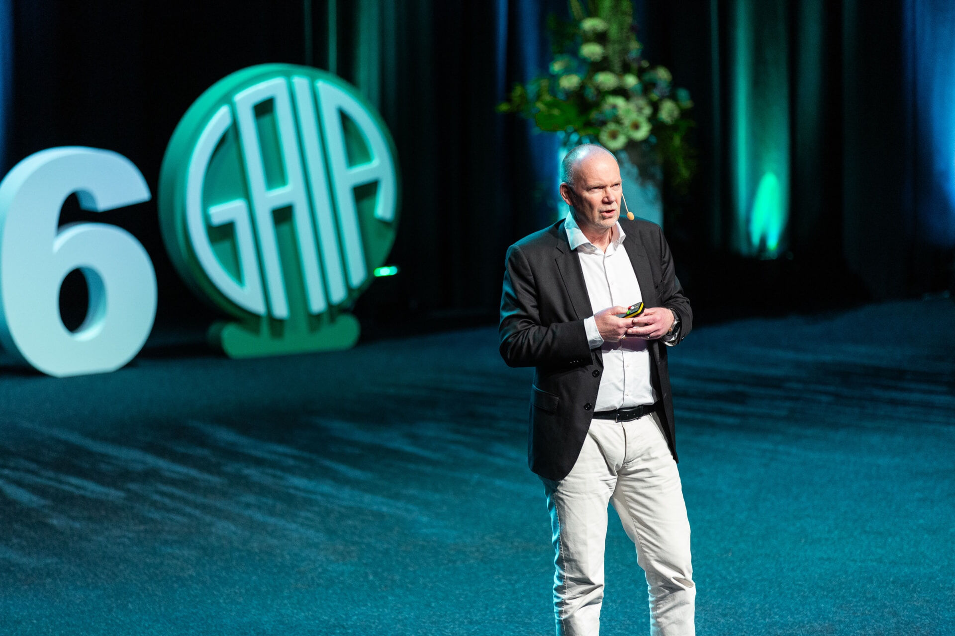 A man speaks on the GAIA Conference stage, an illuminated GAIA logo glowing behind him.